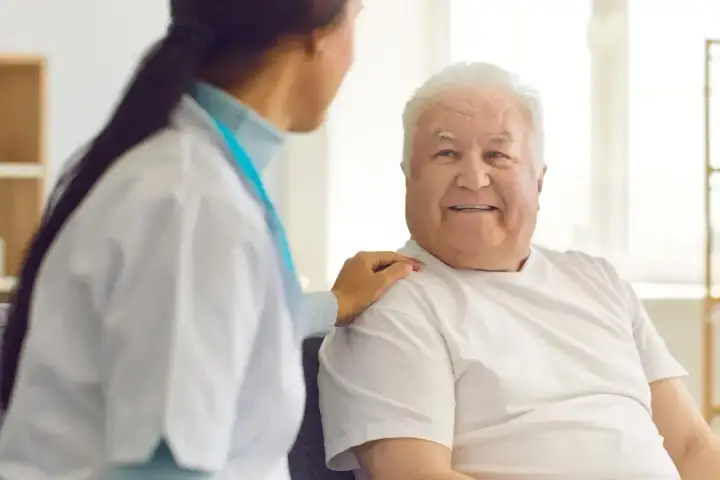 Uśmiechnięty starszy pan z białą czupryną, którego pielęgniarka wspiera ramieniem. To obraz troski i nadziei na świadczenie pielęgnacyjne 2026.
