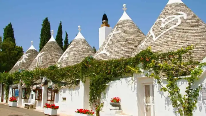 I trulli di Puglia, con i loro tetti conici e le decorazioni bianche, incantano sotto il cielo azzurro, avvolti dal verde rampicante.
