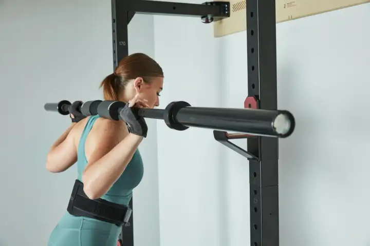 Kobieta w rękawiczkach i pasie treningowym przygotowuje się do przysiadów ze sztangą. Trening push pull legs to jej sposób na siłę.