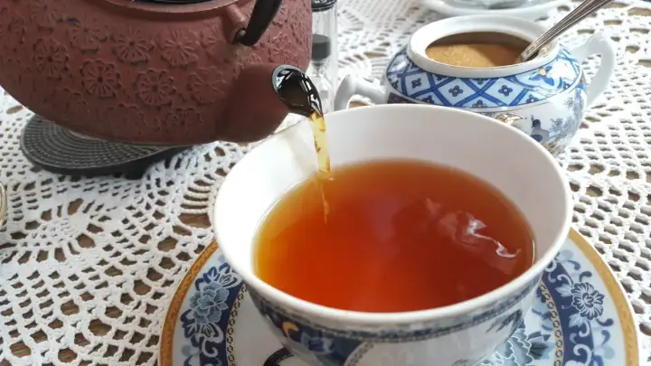 Ciemnobrązowy kolor herbaty w filiżance, nalewanej z ozdobnego czajniczka. Obok cukiernica z brązowym cukrem.