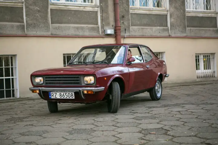 Czerwony fiat 128, klasyk z lat 70., zaparkowany na brukowanym dziedzińcu przed budynkiem.