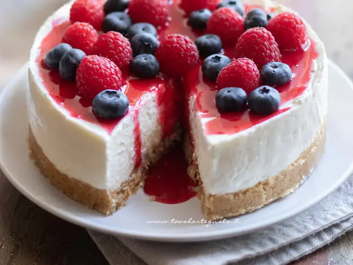 Cheesecake ricetta originale con frutti di bosco freschi e coulis rosso. Una fetta è stata tolta, rivelando la sua consistenza cremosa.