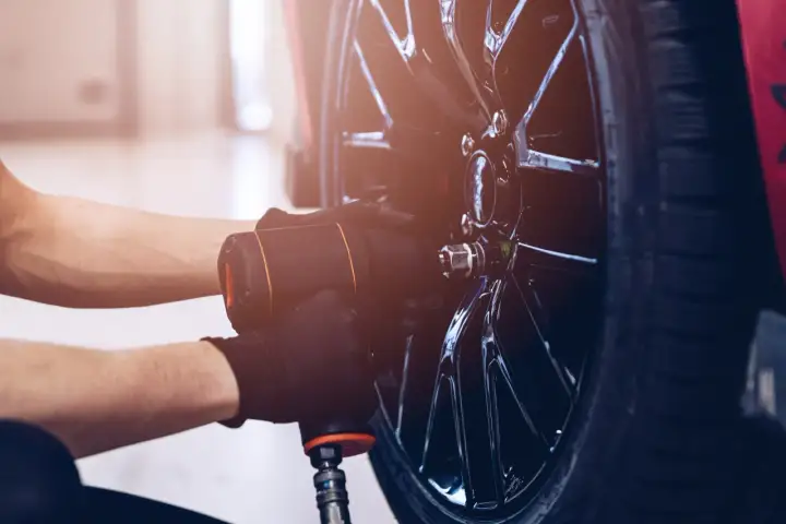Mechanik w czarnych rękawiczkach używa klucza pneumatycznego do odkręcania śrub w feldze, przygotowując się do wymiany jednej opony.