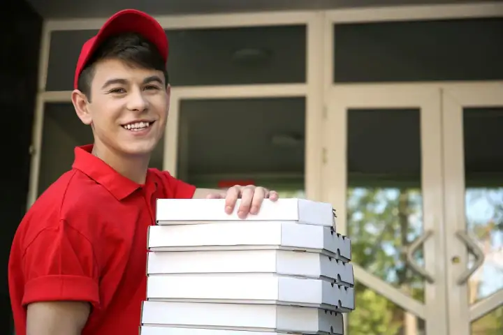 Uśmiechnięty młodociany dostawca pizzy w czerwonej koszulce i czapce trzyma stos pudełek z pizzą przed drzwiami.