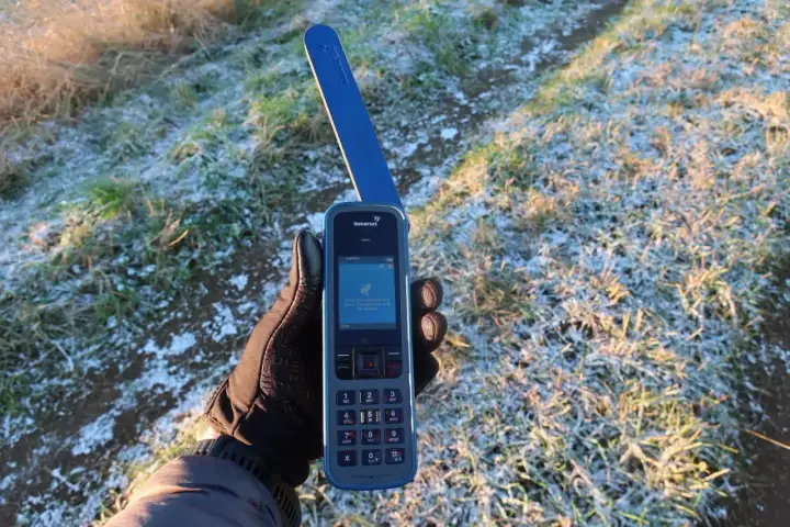 Ręka w rękawiczce trzyma telefon satelitarny Inmarsat IsatPhone, pokazując ekran z informacją o wyszukiwaniu sygnału. Idealne narzędzie, jak sprawdzić prognozę pogody przez telefon satelitarny na wyprawie.