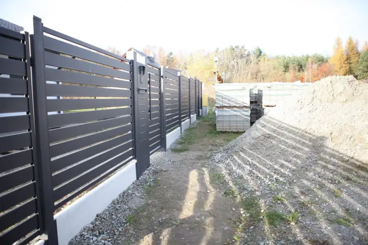Schemat fundamentu pod bramę przesuwną. Widać nowoczesne ogrodzenie, materiały budowlane i hałdę żwiru.