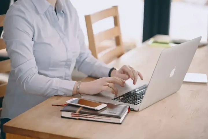 Kobieta w jasnej koszuli pracuje na laptopie, obok leży telefon i notatnik. Dba o swoje ubezpieczenie chorobowe.