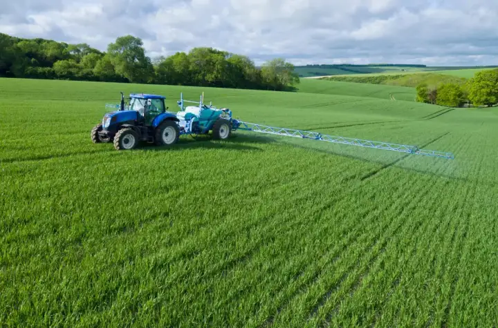 Niebieski traktor z opryskiwaczem na zielonym polu, stosuje środek Chwastox Turbo 340 SL. Dawkowanie jest kluczowe dla skuteczności.