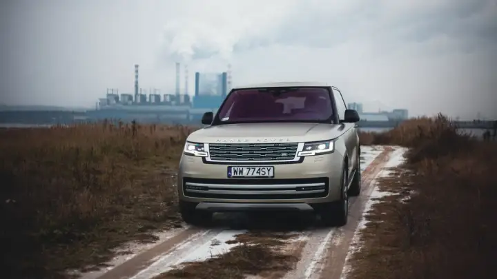 Złoty Range Rover Autobiography na błotnistej drodze, w tle przemysłowy krajobraz z kominami fabrycznymi.