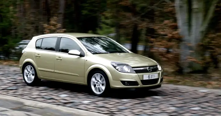 Złoty Opel Astra H 1.7 CDTI w ruchu na brukowanej drodze, otoczony rozmazanym tłem lasu.