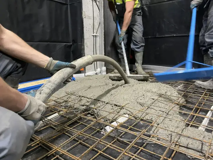Pracownicy wykonują pompowanie betonu na zbrojenie. Beton płynie z węża, a drugi pracownik rozprowadza go łopatą.
