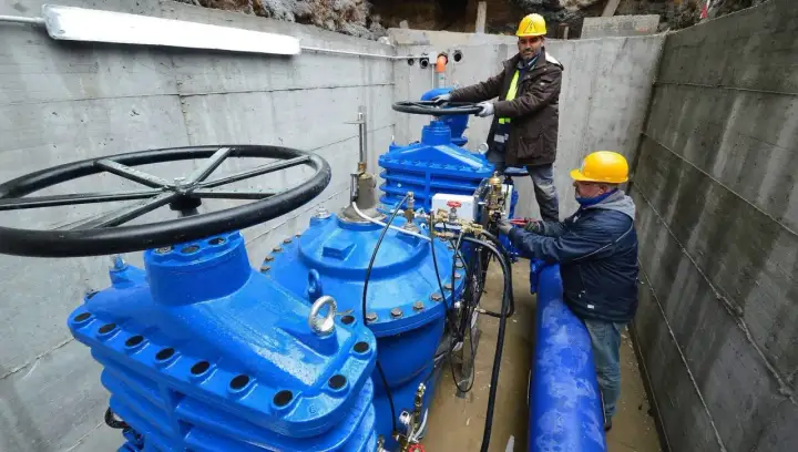 Lavoratori con elmetti gialli operano su grandi valvole blu di un acquedotto. Previste 200 assunzioni per il lavoro acquedotto pugliese.
