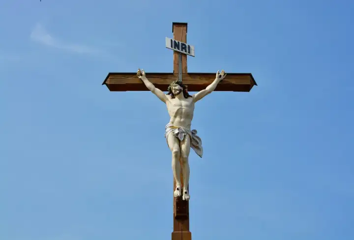 Figurka Jezusa na drewnianym krzyżu z tabliczką INRI na tle błękitnego nieba.
