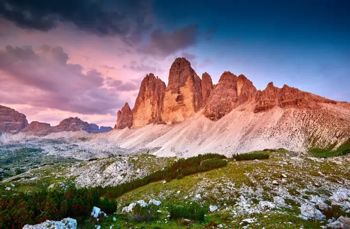 Le maestose cime di Lavaredo illuminate dal tramonto, con un cielo viola e blu che fa da sfondo a un paesaggio alpino mozzafiato.