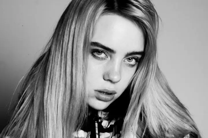 Zbliżenie na twarz Billie Eilish, artystki znanej z unikalnych utworów. Jej spojrzenie jest intensywne, a długie włosy opadają na ramiona.