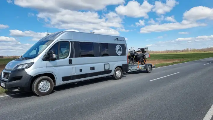 Srebrny kamper z motocyklem na przyczepie jedzie drogą. Przygoda w Szwecji kamperem dopiero się zaczyna.