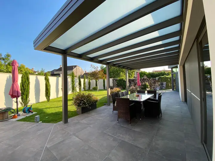 Une belle pergola moderne avec un toit vitré pour couvrir l'espace repas extérieur. Le jardin est bien entretenu, avec une pelouse verte et des cyprès élancés.
