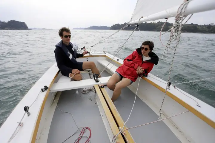 Deux personnes profitent d'une journée ensoleillée sur un voilier dayboat, naviguant sur une mer calme.