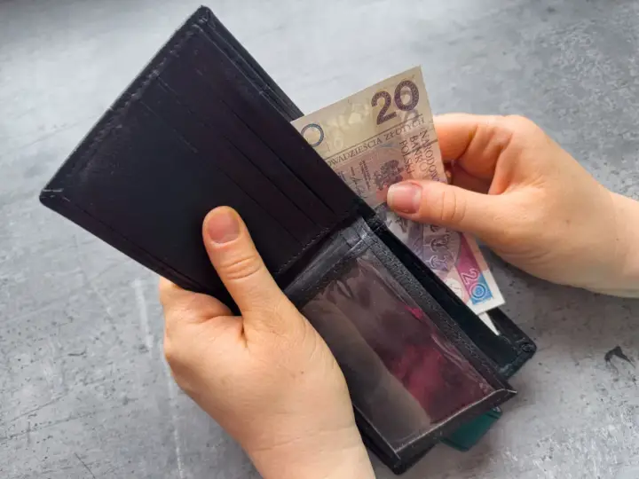 Ręce wkładają banknot 20 zł do portfela. Dowiedz się, komu przysługuje zasiłek dla bezrobotnych.
