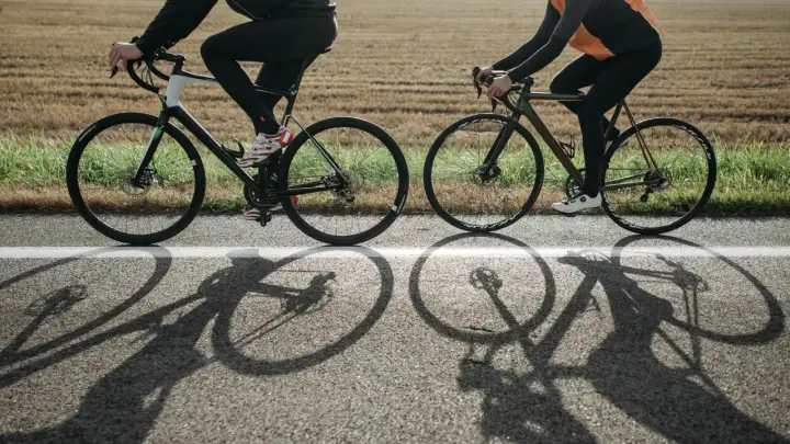 Dwie osoby na rowerach, w kaskach, jadące po drodze. Długie cienie rowerów na asfalcie.
