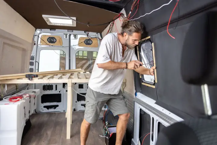 Mężczyzna buduje kamper z osobówki, montując okno. Widać drewniane elementy łóżka i instalację elektryczną.