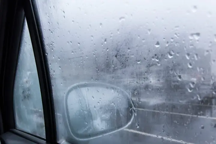 Gotas de lluvia en el cristal del coche, reflejando el exterior y el espejo retrovisor. Una escena que explica porque se empañan los cristales en días húmedos.
