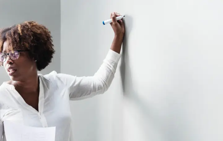 Frau mit Brille und Lockenwicklern hält einen Stift und bietet Nachhilfe an, während sie auf eine weiße Wand blickt.