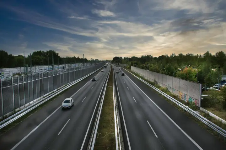 Szeroka autostrada z samochodami w oddali, otoczona zielenią i barierami dźwiękochłonnymi. Jazda autostradą pod pochmurnym niebem.