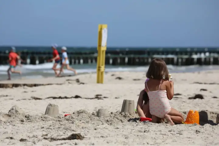 Dzieci budują zamki z piasku na plaży, w tle widać morze i falochrony. Dźwirzyno molo w oddali.
