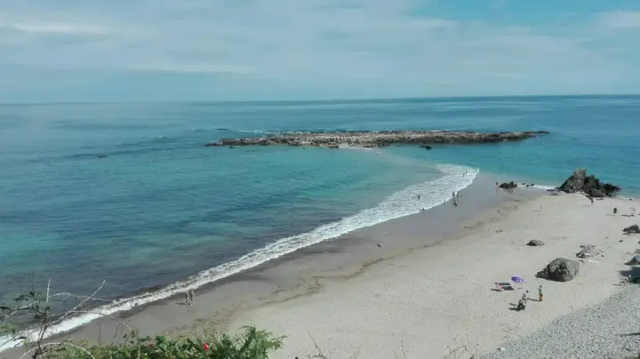 La **playa de Pechón** se extiende con arena dorada y aguas turquesas. Unas rocas emergen del mar, mientras algunas personas disfrutan del sol y el mar.