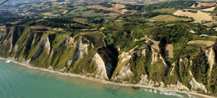 Scogliere a picco sul mare turchese a Gabicce Mare, Marche. Campi coltivati e vegetazione rigogliosa adornano le colline retrostanti.