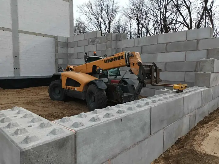 Żółty ładowacz teleskopowy Liebherr pracuje przy budowie muru oporowego z betonowych bloków.