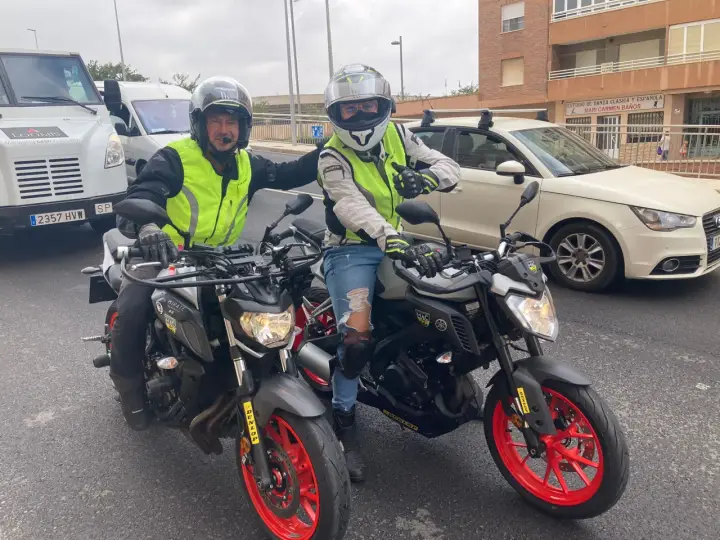 Dos motoristas con chalecos reflectantes y cascos posan con sus motos. Uno hace un gesto de "pulgar arriba". ¡Listos para la aventura en su a1 de moto!