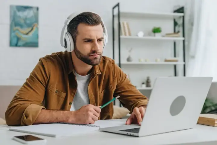 Mann mit Kopfhörern lernt online, nutzt Laptop und Notizbuch. Er wirkt konzentriert, während er mit einem Stift tippt. Ein typisches Bild für das heutige "Home-Office"-Zeitalter, das viele Anglizismen mit sich bringt.