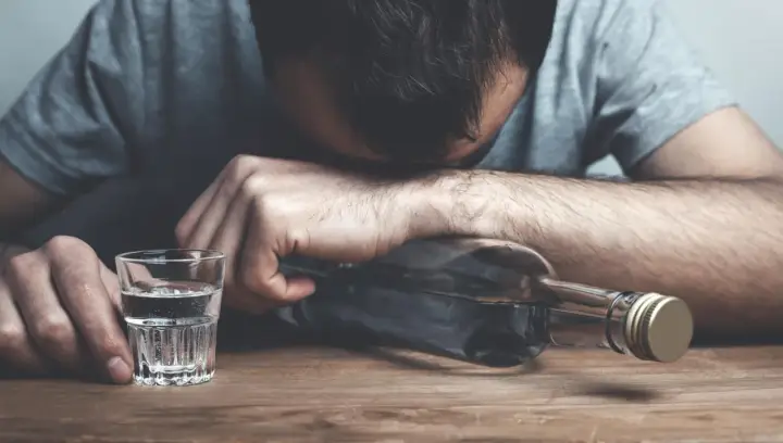 Mężczyzna z głową opartą o butelkę, obok kieliszek z alkoholem. Wygląda na to, że doświadcza zatrucia alkoholowego.