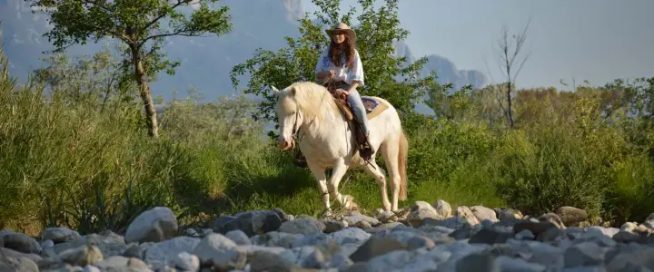 Giovane donna con cappello da cowboy e cavallo bianco durante una passeggiata a cavallo in Campania, tra rocce e vegetazione rigogliosa.