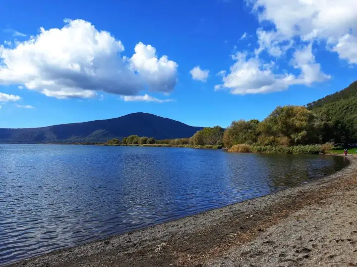 Spiaggia ghiaiosa e acque calme del lago di Vico, con montagne verdi e cielo azzurro punteggiato di nuvole bianche.