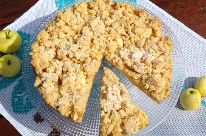 Szarlotka na kruchym cieście z kruszonką, obok jabłka, na talerzu.