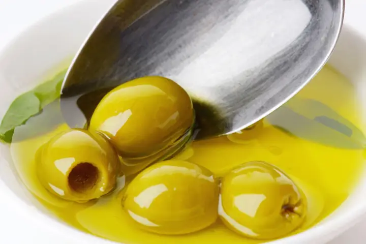 Kilka zielonych oliwek w złocistej oliwie, łyżka zanurzona w płynie. Oliwa z oliwek kcal - zdrowie i smak.