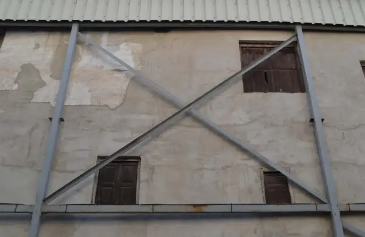 Estructura metálica en forma de X que refuerza una pared de concreto con ventanas de madera. La riostra, en este caso, es un elemento de soporte crucial.