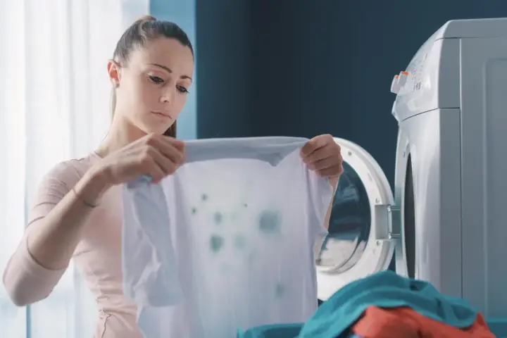 Zmartwiona kobieta trzyma białą koszulkę z zielonymi plamami, która wygląda jak zafarbowane ubrania. Obok stoi otwarta pralka.
