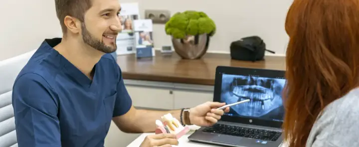 Dentysta omawia z pacjentką plan leczenia, pokazując na ekranie zdjęcie rentgenowskie szczęki i model zębów, wyjaśniając znaczenie czyszczenia dziąseł.