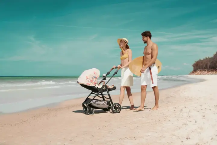 Para spaceruje po plaży z wózkiem Kinderkraft Askoy. Mężczyzna niesie deskę surfingową.