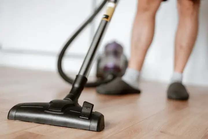 Odkurzacz na drewnianej podłodze, część nóg w kapciach i skarpetkach. To jeden z obowiązków domowych po angielsku – "vacuuming".