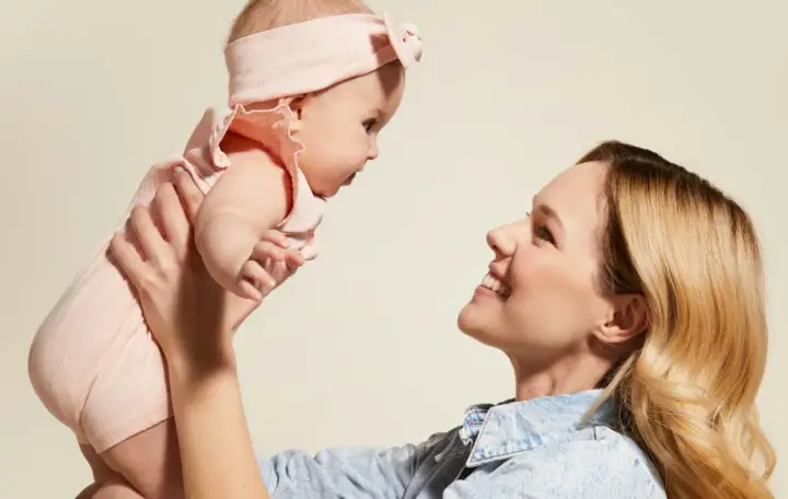 Mama podnosi dziecko w różowym body i opasce. Idealny przykład, jak ubierać noworodka latem – lekko i wygodnie.