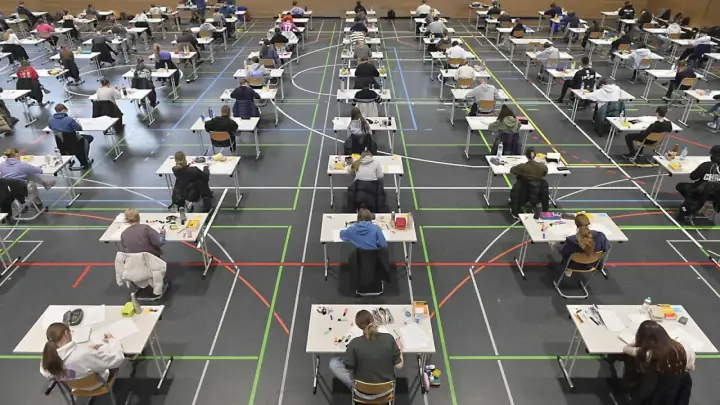 Viele Schüler sitzen in einer Sporthalle an Tischen, um zum Abitur zu schreiben.