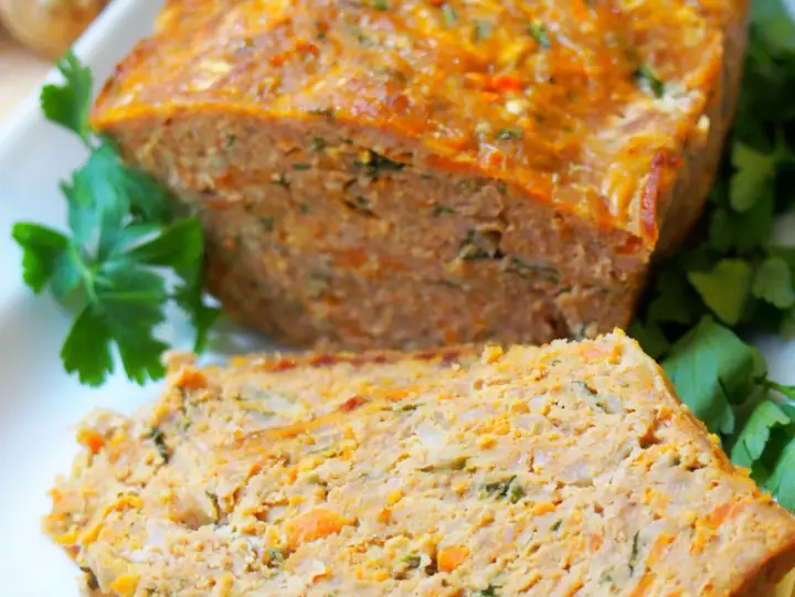 Pyszna pieczeń z mięsa mielonego, pokrojona w plastry, z widocznymi kawałkami marchewki i ziół, udekorowana natką pietruszki.