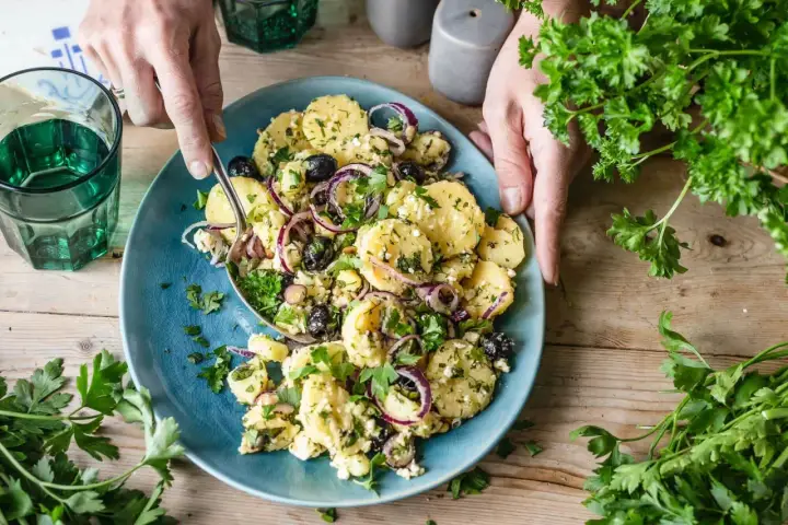 Ein köstlicher mediterraner Kartoffelsalat mit Oliven, roten Zwiebeln und Feta auf einem blauen Teller, garniert mit frischer Petersilie.