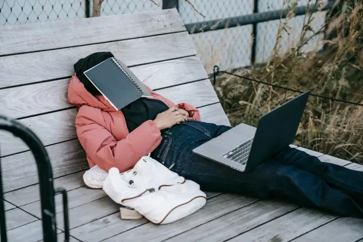Osoba w różowej kurtce leży na ławce, z zeszytem na twarzy. Obok leży laptop i plecak. Czas na odpoczynek i dodanie nowych pomysłów.