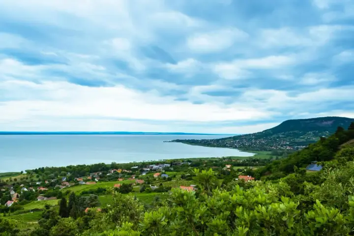 Widok na Balaton z góry, z zielonymi wzgórzami i domkami. W oddali widać jezioro i plażę.